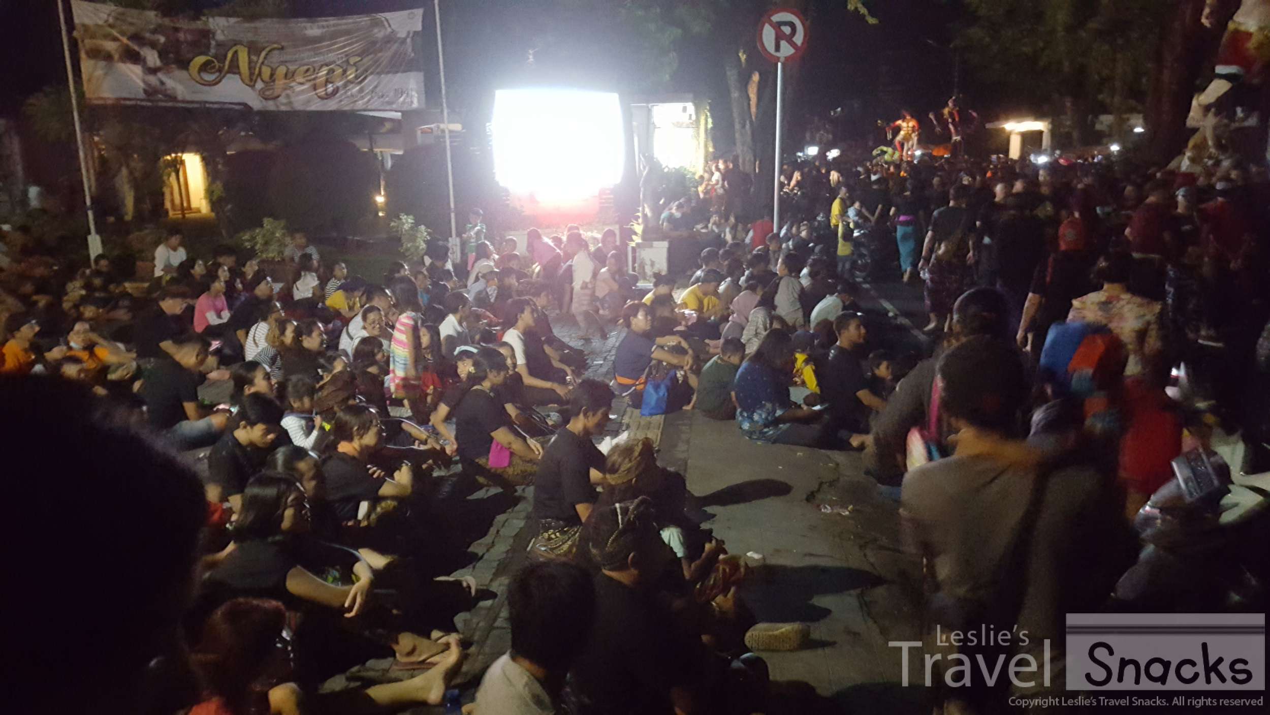 In front of the Inna Bali Heritage Hotel, people sit and watch as the procession waits for its turn to enter the main roundabout.